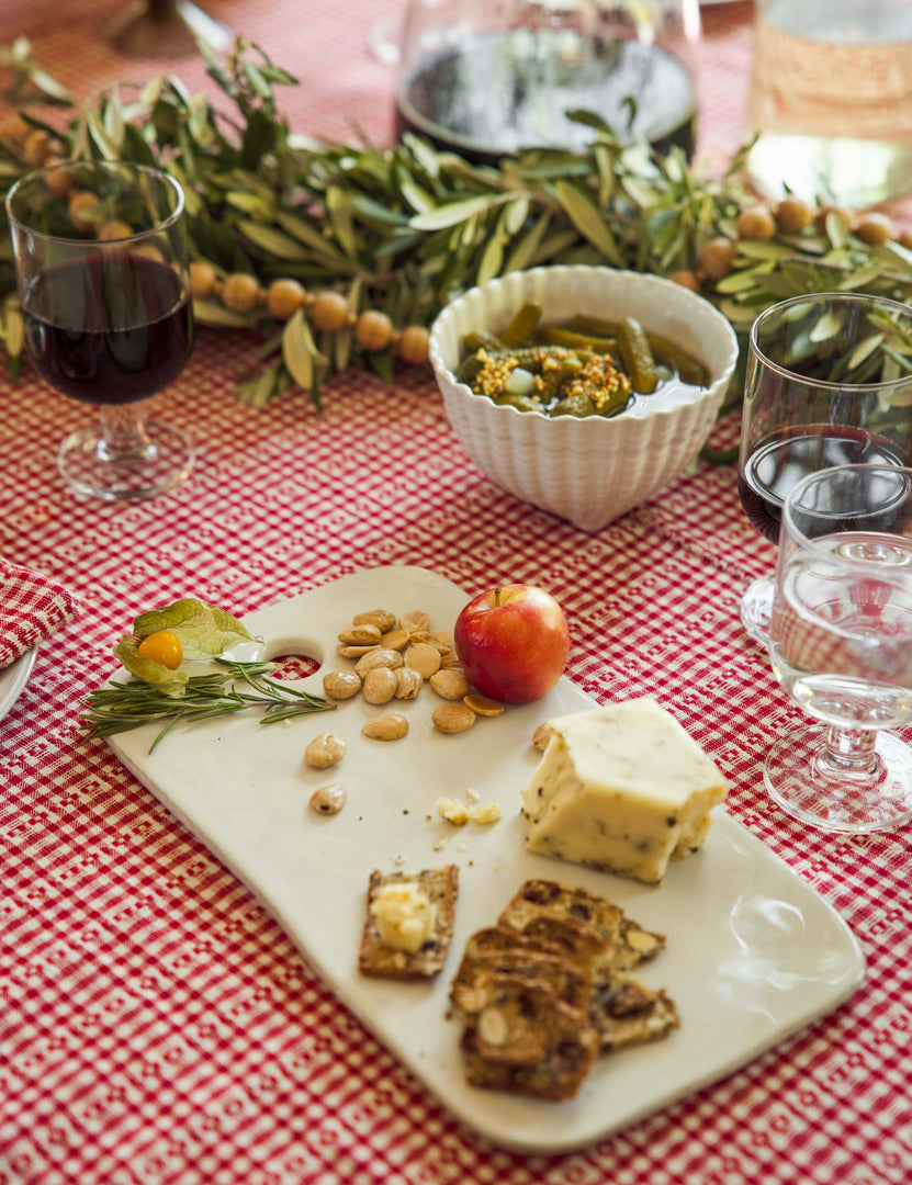 | The Aparte white footed bowl with shell inspired design by Costa Nova sits on a red plaid tablecloth in a dining room with a marble cheese board and clear wine glasses