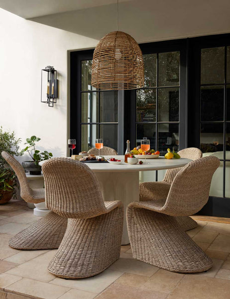 #color::natural | The Manila wicker weave beige indoor and outdoor dining chair sits in a covered outdoor dining space beneath a natural jute chandelier surrounding an ivory circular dining table