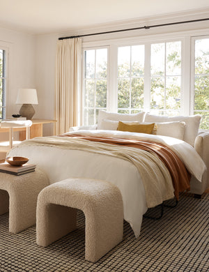 Two Tate Cream Boucle stools sit at the end of a bed on a gridded rug with a wooden bowl and a stack of books on one of them