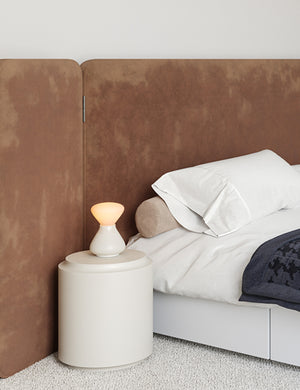 The Reflection Noma white table lamp by Tala sits in a bedroom atop a white nightstand with a brown velvet framed bed to its right