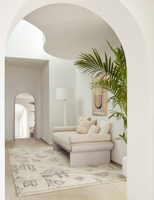 The Shashenka silver arched floor mirror sits in a sculptural space with a cream cushioned bench, a plush patterend rug, and a white lamp with a ribbed base